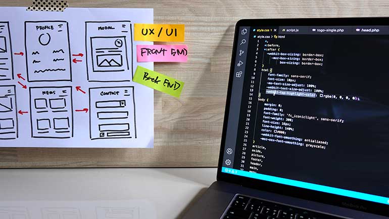 Coding can be seen on laptop screen and UI on a paper for website development